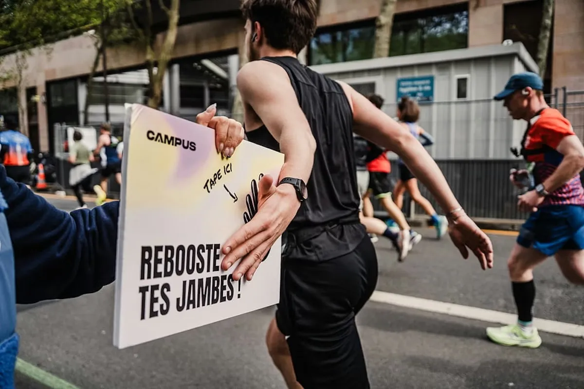 At the edge of a race course, a hand holds out a Campus sign that says « tape ici, reboost tes jambes ». A runner slaps it as he passes.