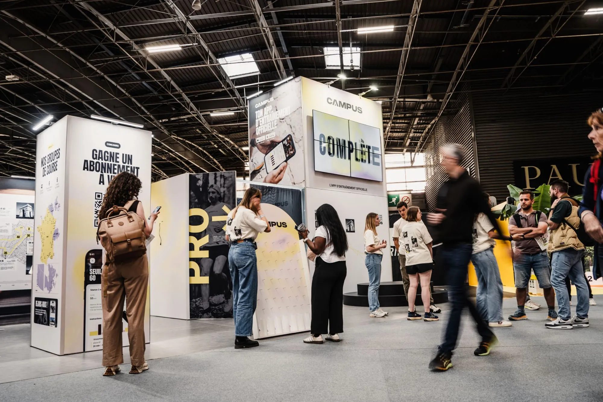 A Campus booth in an exhibition hall. Two-meter totems carry blown-up screens of the app, a threshold session and a map of running groups. A wall of screens displays the word complète. Visitors stop in front.